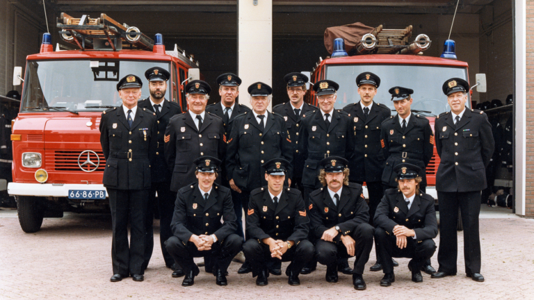 Groepsfoto van brandweerlieden in uniform voor twee rode brandweerwagens, geposeerd op een binnenplaats bij een brandweerkazerne.