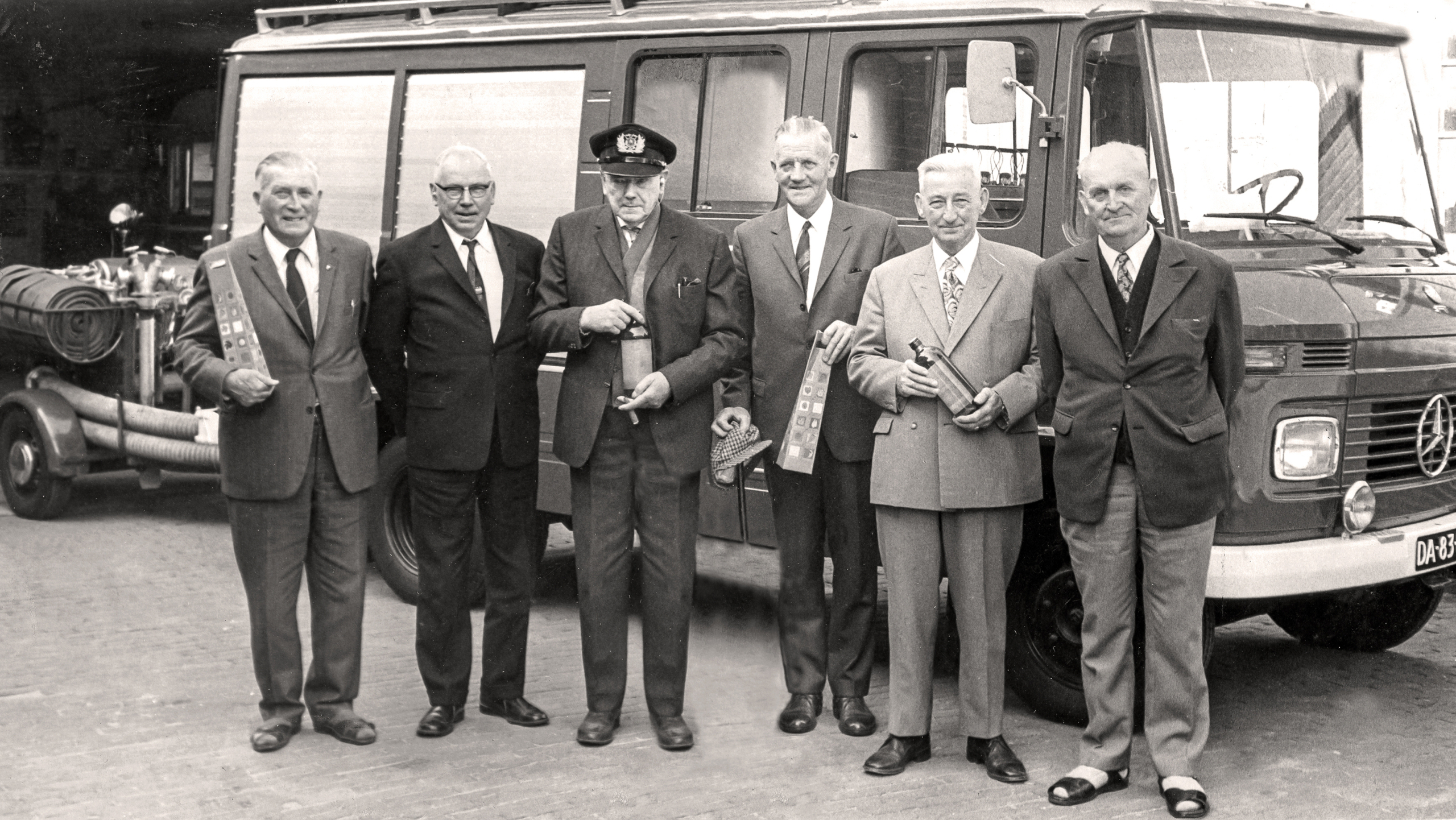 Een zwart-witfoto van zes mannen in nette pakken die poseren voor een brandweerwagen bij een brandweerkazerne. Ze houden allemaal voorwerpen vast, zoals flessen en andere voorwerpen. Boven de ingang van de kazerne is het woord 'BRANDWEER' zichtbaar.