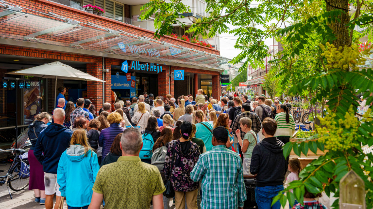 Een grote groep mensen staat in de rij voor de ingang van een Albert Heijn supermarkt, omringd door groene bomen en fietsen.
