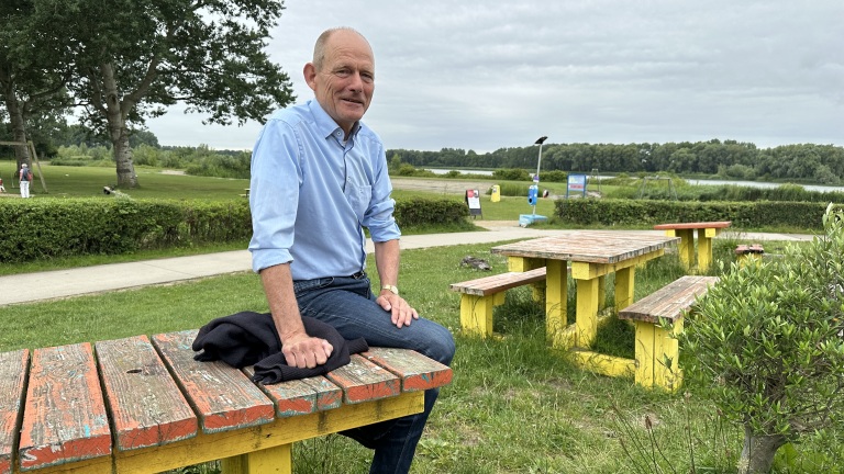 Een man zit op een kleurrijke picknicktafel in een park met gras, bomen en een pad op de achtergrond.