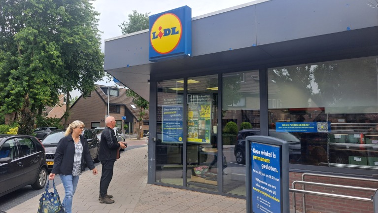 Vooraanzicht van een gesloten Lidl-winkel met borden op de deur die de sluiting aankondigen, naast een voorbijlopende vrouw en een man die richting de winkel kijkt, in een straat met parkeergelegenheid en groene bomen.