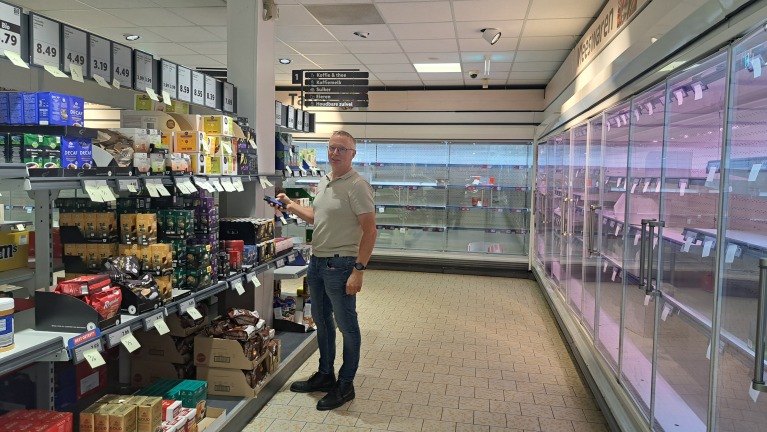 Een man staat in een supermarkt bij een schap met verschillende soorten koffie; naast hem zijn lege koelschappen te zien.