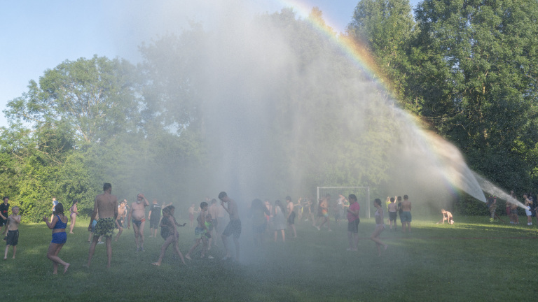 Groep mensen speelt in een park onder een waterstraal, met een regenboog zichtbaar in de mist.