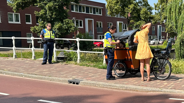 Twee politieagenten controleren een vrouw met een bakfiets op een zonnige weg naast een kanaal met bomen en huizen op de achtergrond.