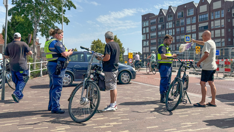 Twee handhavers spreken met mannen bij hun fietsen op een zonnige dag; op de achtergrond zijn gebouwen en een paar passerende fietsers te zien.
