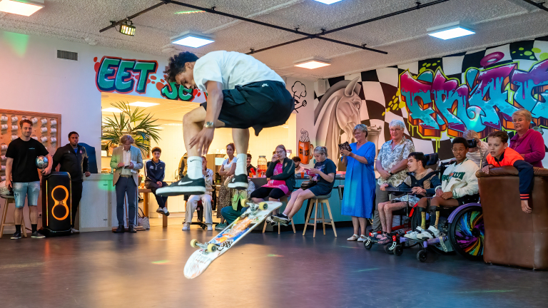 Een skateboarder voert een truc uit in een binnenruimte met kleurrijke graffiti, terwijl een groep mensen toekijkt, inclusief kinderen in rolstoelen.