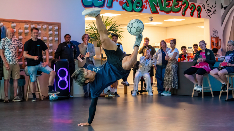 Een persoon voert een freestyle voetbaltruc uit op één hand, terwijl een groep mensen toekijkt in een interieur met graffiti op de muur.