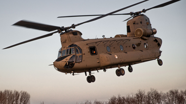 Een militaire helikopter, specifiek een Boeing CH-47 Chinook, vliegt boven een bosrijk gebied tegen een heldere lucht.