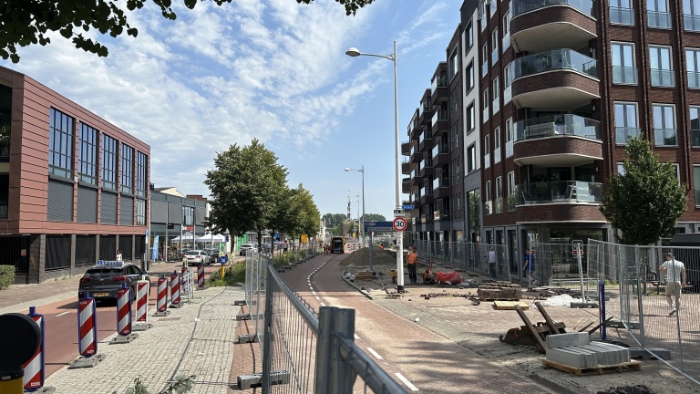 Wegwerkzaamheden aan een straat met moderne gebouwen aan beide zijden, borden voor snelheidsbeperking en diverse bouwwerkzaamheden op de achtergrond, onder een blauwe lucht.