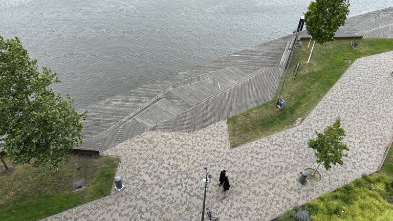 Een bovenaanzicht van een pad langs een waterkant met houten vlonders, grasvelden en bomen, waar enkele mensen wandelen.