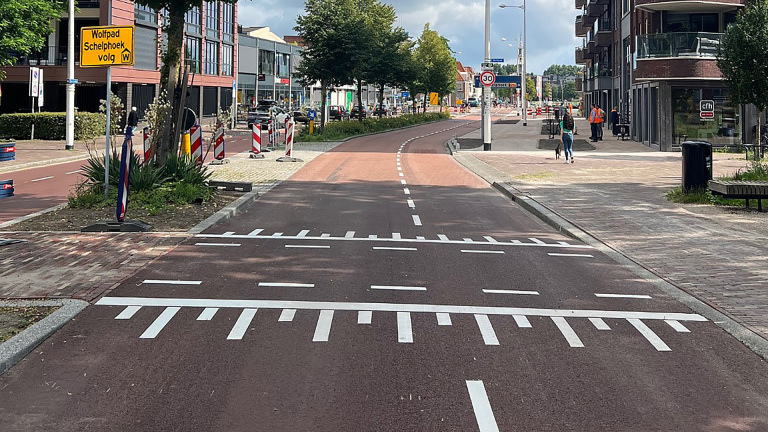 Straatbeeld met een gescheiden fietspad en bord met tekst 'Wolfpad Schelphoek volg', omringd door stedelijke bebouwing en verkeersborden.