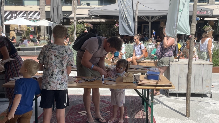 Verschillende mensen, inclusief kinderen, doen mee aan een creatieve workshop aan een tafel buiten op een plein bij een coffeeshop en lunchcafé. Een volwassene helpt een kind met verf op haar gezicht bij het knutselen met papier en lijm. Om hen heen zijn andere mensen te zien in een zonovergoten omgeving.