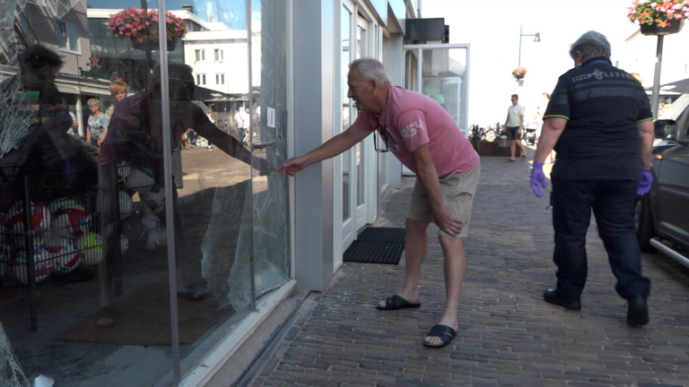 Een oudere man in een roze T-shirt inspecteert een gebroken glazen winkelruit, terwijl een persoon in uniform met paarse handschoenen erbij staat op een zonnige dag in een winkelstraat.