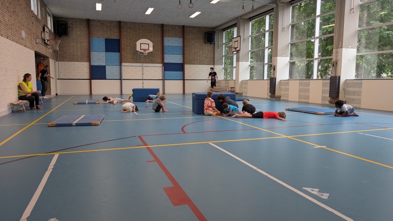 Kinderen spelen in een gymzaal, liggend op de grond of op matten, terwijl enkele volwassenen toekijken.
