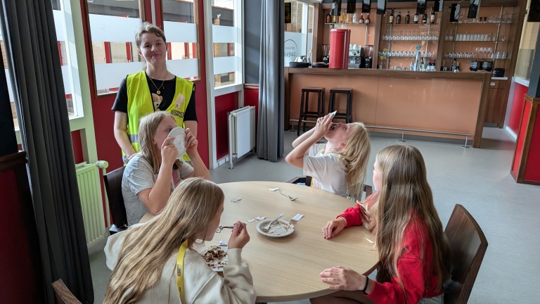 Een groep kinderen zit rond een tafel in een eetruimte, waarbij sommigen spelen met lege borden, terwijl een volwassene in een geel hesje toekijkt.