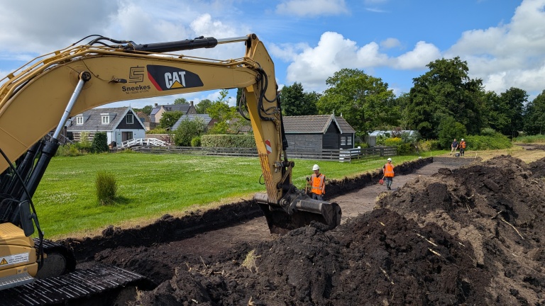 Graafmachine met CAT-logo graaft een greppel terwijl arbeiders in oranje veiligheidshesjes en helmen toezicht houden, met huizen en groen in de achtergrond op een bewolkte dag.