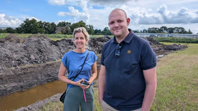 Een man en een vrouw staan buiten voor een bouwplaats met een sloot op de achtergrond. Er liggen hopen aarde en gras om hen heen en blauwe lucht met wolken erboven.