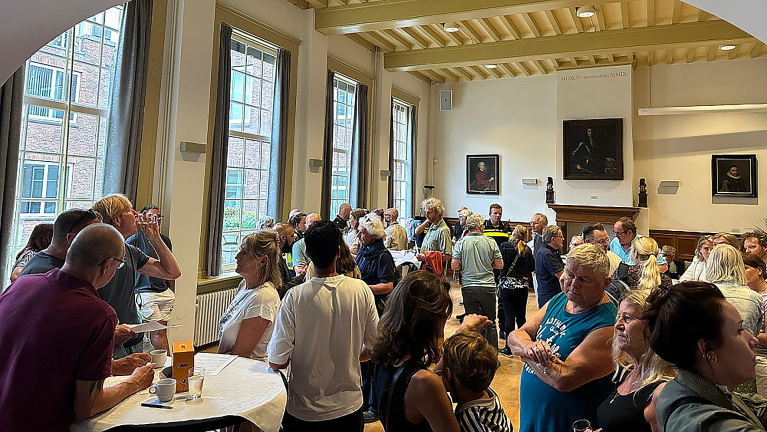 Een groep mensen verzameld in een kamer met hoge ramen en schilderijen aan de muren, bezig met sociale interacties en drinken.