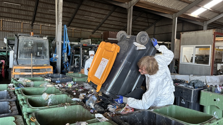 Een persoon in een beschermend pak kiept een grote zwarte container met een oranje deksel in een rij groene afvalbakken in een recyclingcentrum.