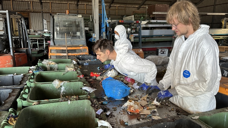 Mensen in witte pakken en handschoenen, bezig met afvalsortering in een industriële omgeving.