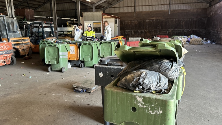 Binnen een loods staan verschillende groene en oranje afvalcontainers. Op de achtergrond zijn werknemers te zien in werk- en veiligheidskleding die bezig zijn met het sorteren van afval. Aan de rechterkant zijn enkele zakken met afval zichtbaar in de voorgrond.