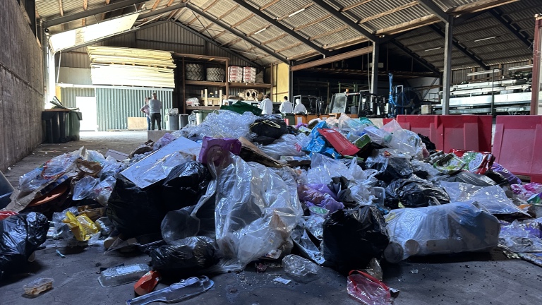 Een grote stapel afval, bestaande uit plastic zakken en verpakkingen, ligt op de vloer van een loods. Op de achtergrond zijn twee mensen te zien die met elkaar praten, naast opslagrekken en apparatuur.