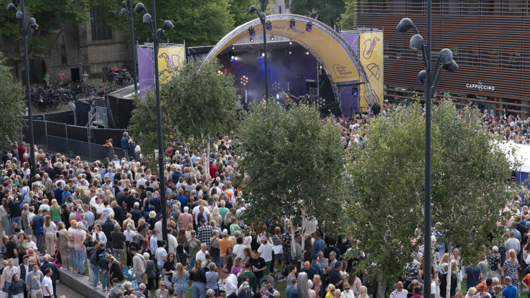 Grote menigte verzameld voor een openluchtconcert bij een groot podium met verlichting en omliggende bomen, met op de achtergrond een gebouw met het opschrift 'Cappuccino'.