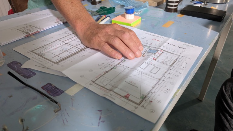 Een hand rust op een gedetailleerde bouwtekening, omringd door tekenspullen, op een tafel met een blauwe verflaag.