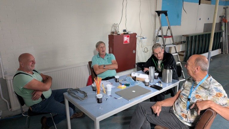 Vier oudere mannen zitten rond een tafel in een ongeorganiseerde kamer, met koffiekopjes en een laptop voor hen op tafel. Er zijn een ladder en diverse bouwmaterialen op de achtergrond zichtbaar.