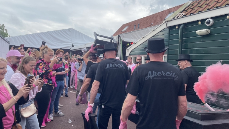 Groep mensen bij een evenement, met sommigen in zwarte hoeden en t-shirts met 'Akercity', terwijl een persoon iets omhoog tilt. Omstanders maken foto's en een roze rookwolk is te zien.