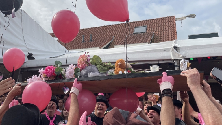 Groep mensen verzameld onder een houten plank versierd met bloemen, ballonnen en knuffels, opgehouden door personen met roze handschoenen.