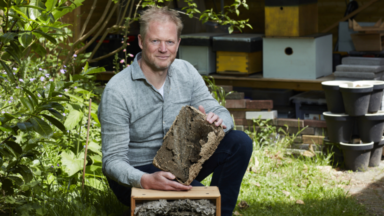 Een man zit gehurkt in een tuin en houdt een stuk bijenraat vast. Op de achtergrond zijn een bijenkast en verschillende planten te zien.