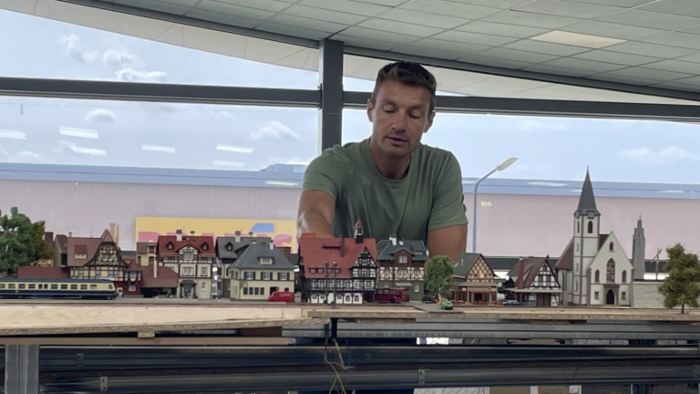 Een man in een groene T-shirt observeert een modeltrein en miniatuurstation in een grote hal met een glazen wand.