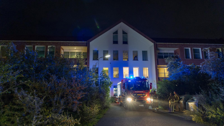 Een brandweerwagen met knipperende lichten staat 's nachts voor een groot gebouw met meerdere verdiepingen, omgeven door struiken. Brandweerlieden staan naast de wagen.