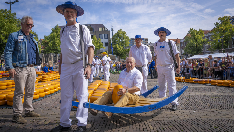 Mannen in witte kleding en blauwe hoeden dragen een kaasbrancard met ronde kazen op een marktplein, omringd door toeschouwers en gebouwen.