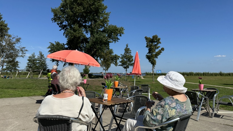 Twee oudere vrouwen zitten op een terras met tafels en stoelen, onder een roze parasol op een zonnige dag, met uitzicht op een groene omgeving en blauwe lucht.