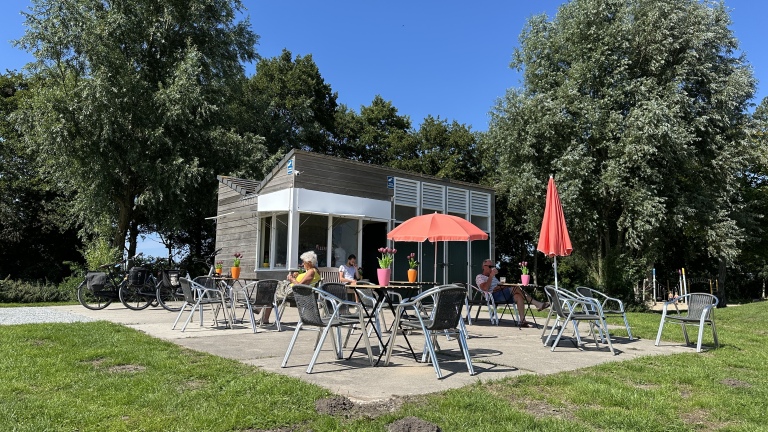 Een klein café met een terras met enkele tafels en stoelen onder rode parasols, waar mensen genieten van drankjes in een groene omgeving met grote bomen.