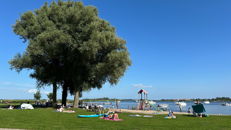 Mensen ontspannen op een grasveld naast een meer met enkele boten en een speeltoestel in de buurt, onder een heldere blauwe hemel.