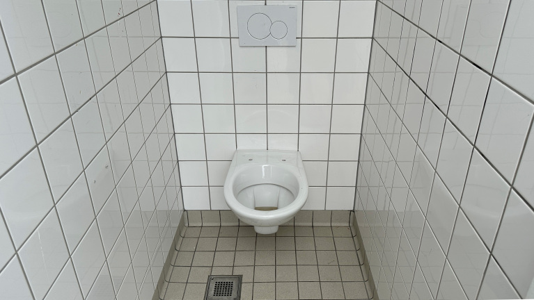 Een wit, hangend toilet in een badkamercabine met witte metrotegels op de muren en grijze tegels op de vloer.