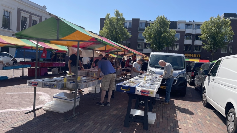 Markt met kleurrijke kraampjes waar mensen door bakken met boeken of tijdschriften bladeren, met op de achtergrond een gebouw en enkele geparkeerde bestelwagens.