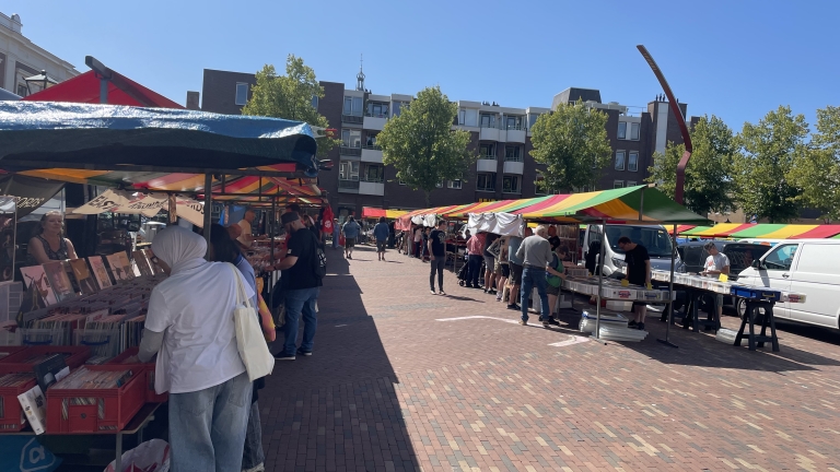 Straatmarkt met kleurrijke kraampjes en mensen die platen bekijken; op de achtergrond staan gebouwen en bomen.