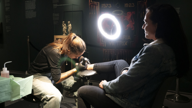 Een tatoeΓ«erder bezig met het tatoeΓ«ren van een vrouw in een donkere kamer, met een heldere ringlamp die het gebied verlicht.