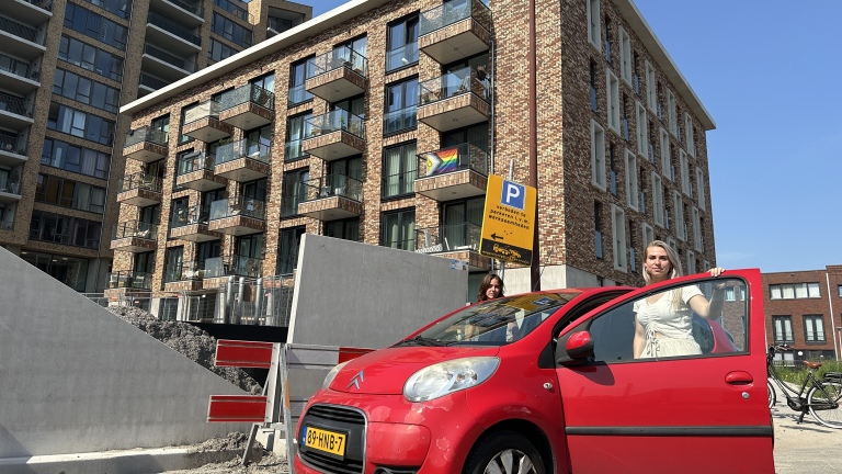 Twee mensen staan bij een geopende rode auto voor een hoog gebouw met balkons, terwijl een regenboogvlag zichtbaar is. Op de achtergrond is een parkeerbord te zien en er staat een fiets geparkeerd naast een stapel zand en betonnen constructies.