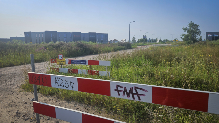 Een bouwterrein met borden en rode-witte afzettingen, waarop teksten zoals "Holland op zijn smalst" en graffiti te zien zijn. Op de achtergrond zijn industriële gebouwen en straatlantaarns zichtbaar, omgeven door hoog gras.