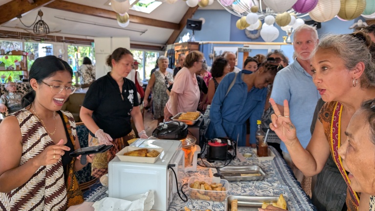 Mensen staan in een rij bij een eettafel met snacks, terwijl een vrouw iets aan een andere vrouw uitlegt in een drukbezocht interieur met decoratieve lampionnen.