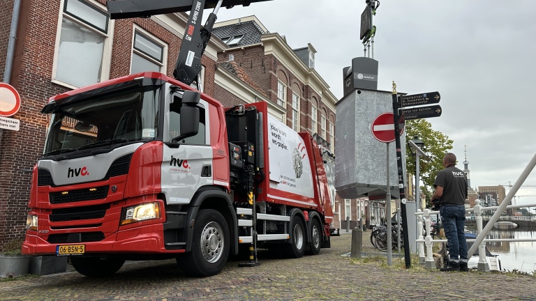 HVC-vuilniswagen met kraan tilt ondergrondse afvalcontainer naast kanaal, met medewerker en verkeersborden op de kade