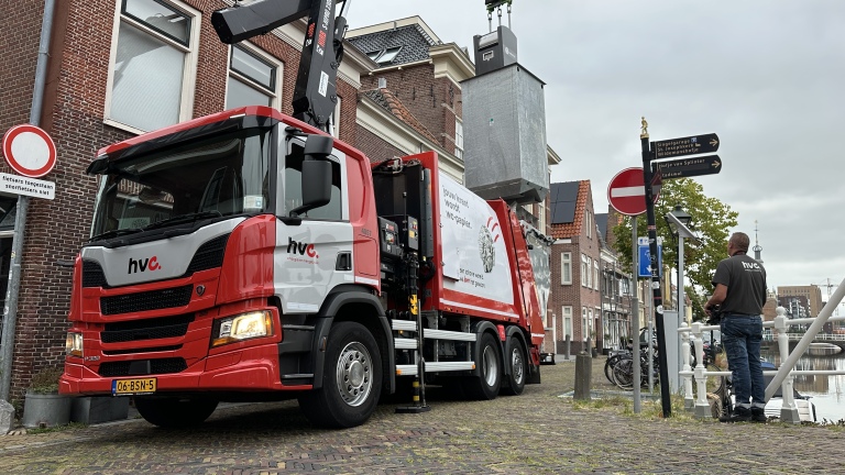 Rode HVC-vrachtwagen met kraan hijst grijze container in smalle straat langs gracht, medewerker staat ernaast