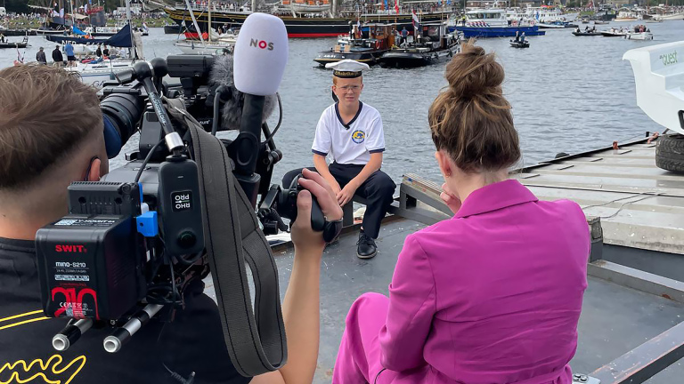 Een jongen in een matrozenkostuum wordt geïnterviewd en gefilmd aan de waterkant, met boten en een drukke haven op de achtergrond.