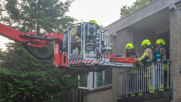 Brandweerlieden in gele helmen en beschermende kleding tillen een brancard naar een platform met een hijskraan aan de zijkant van een gebouw.