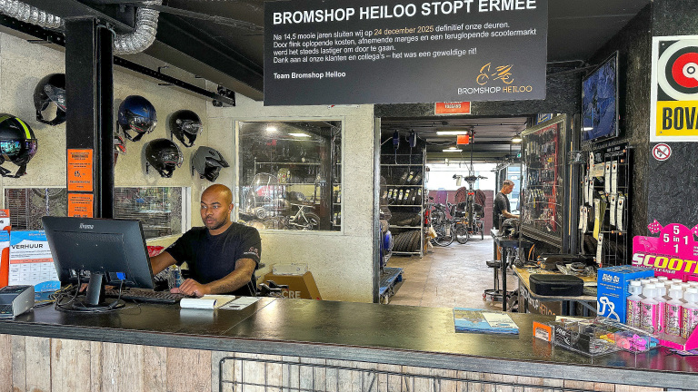 Man achter de balie in een scooterwinkel, met helmen aan de muur en een bord met de tekst over de sluiting van de winkel Bromshop Heiloo.